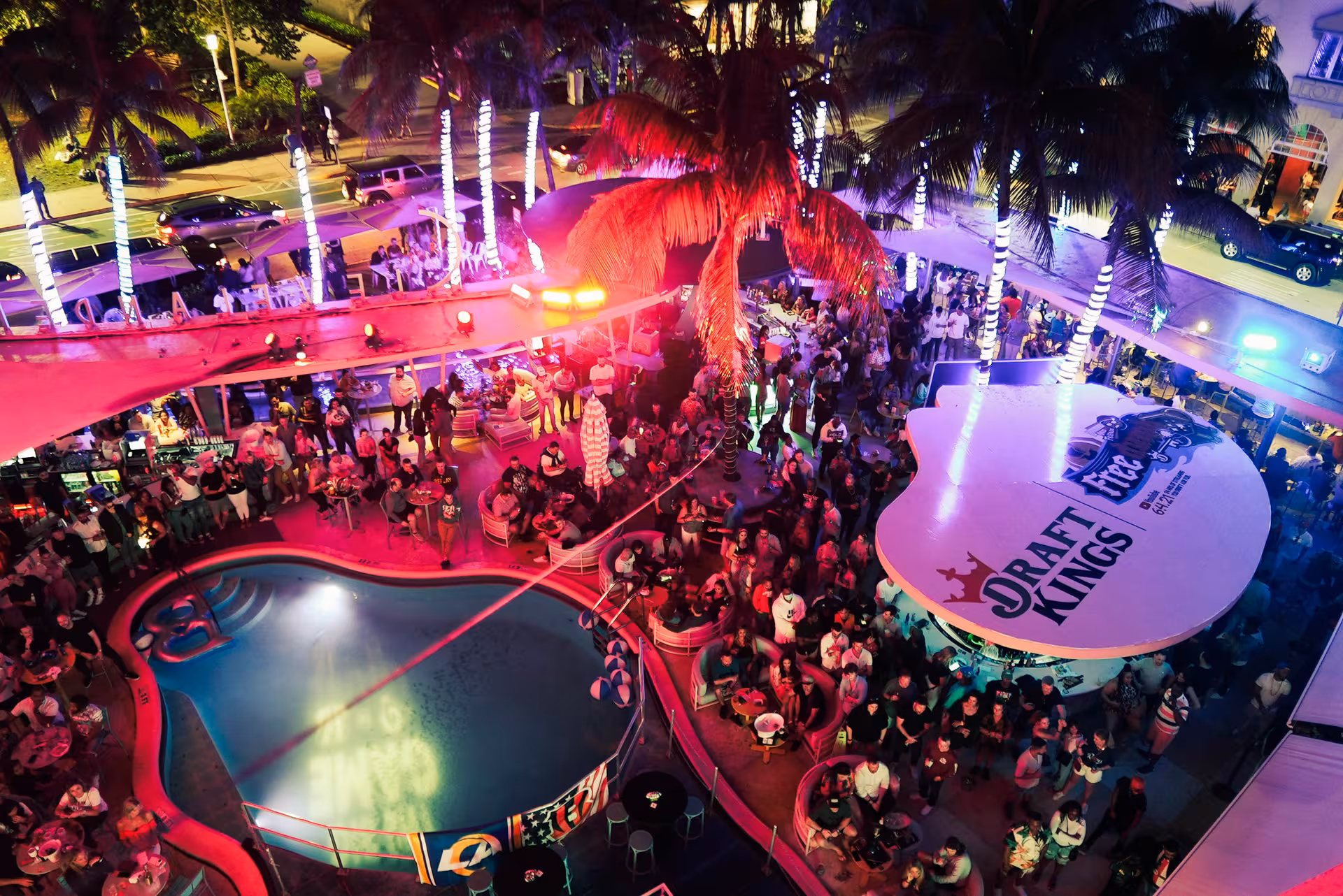 A crowded pool area at night with a Draft Kings sign and lit palm trees.