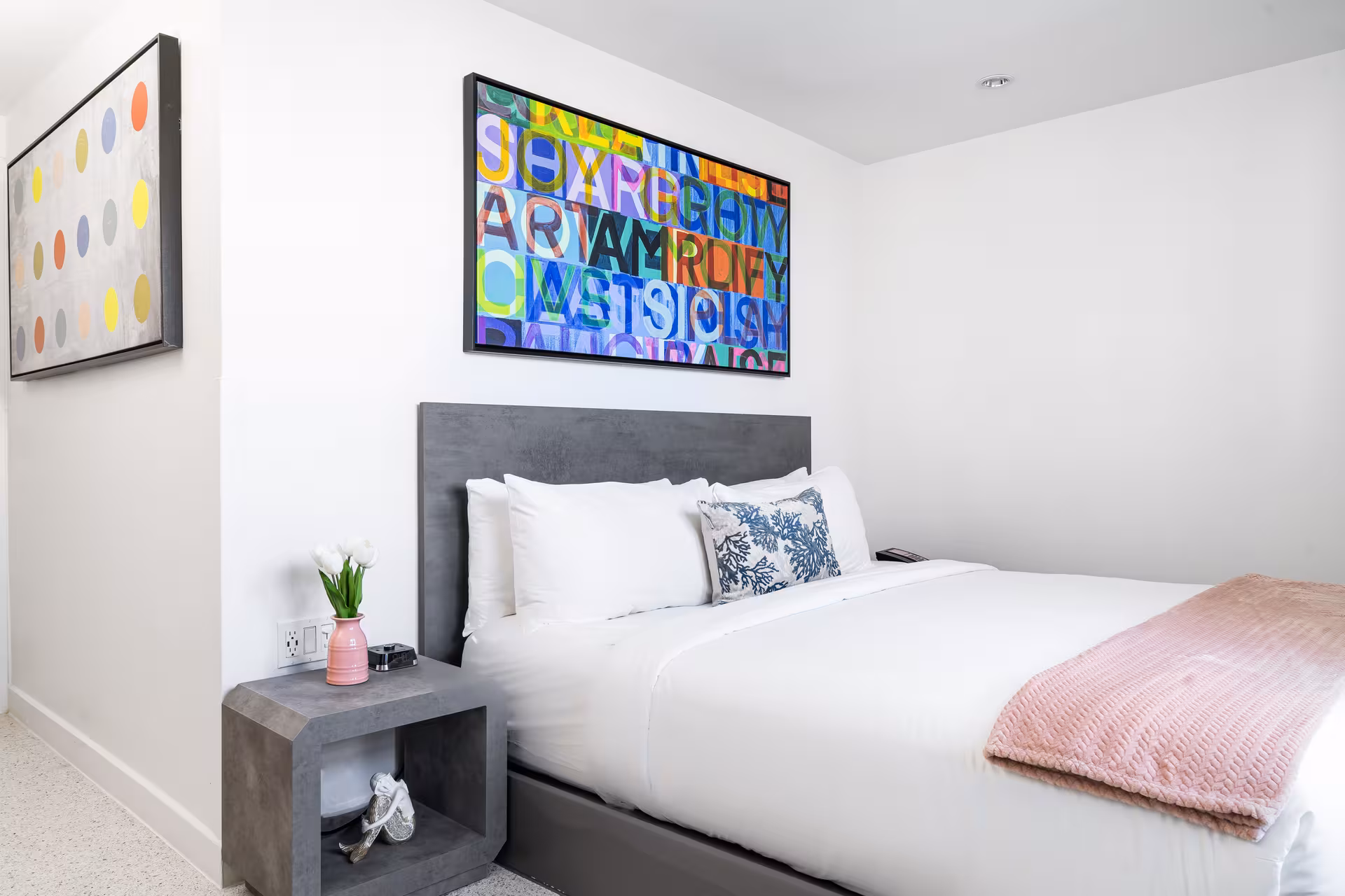 A neatly made bed with white linens and a gray headboard in a room with colorful artwork and a pink vase on a nightstand.