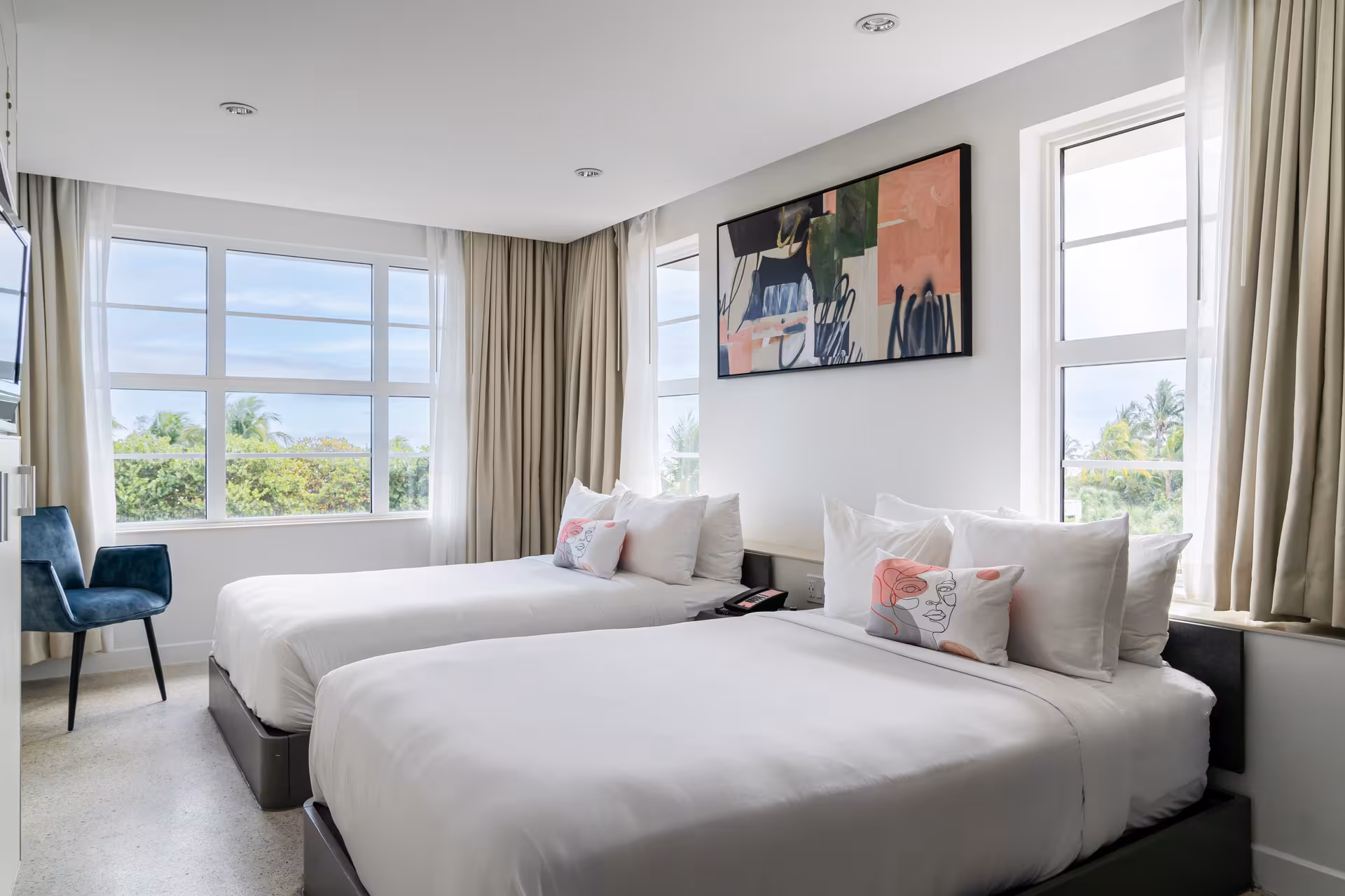 A hotel room with two twin beds, large windows, and a blue chair.