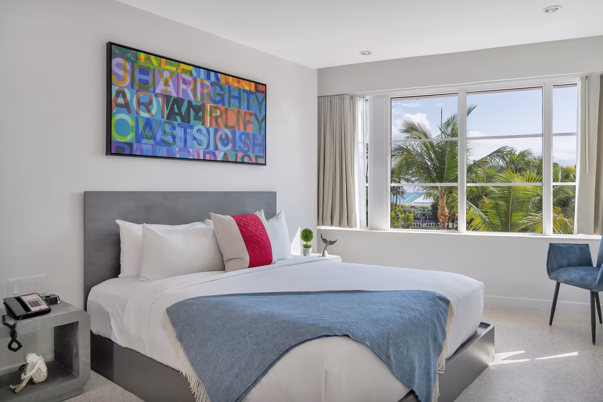 A wide-angle view of a hotel room with a king-sized bed, a large window overlooking palm trees, and modern artwork on the wall.