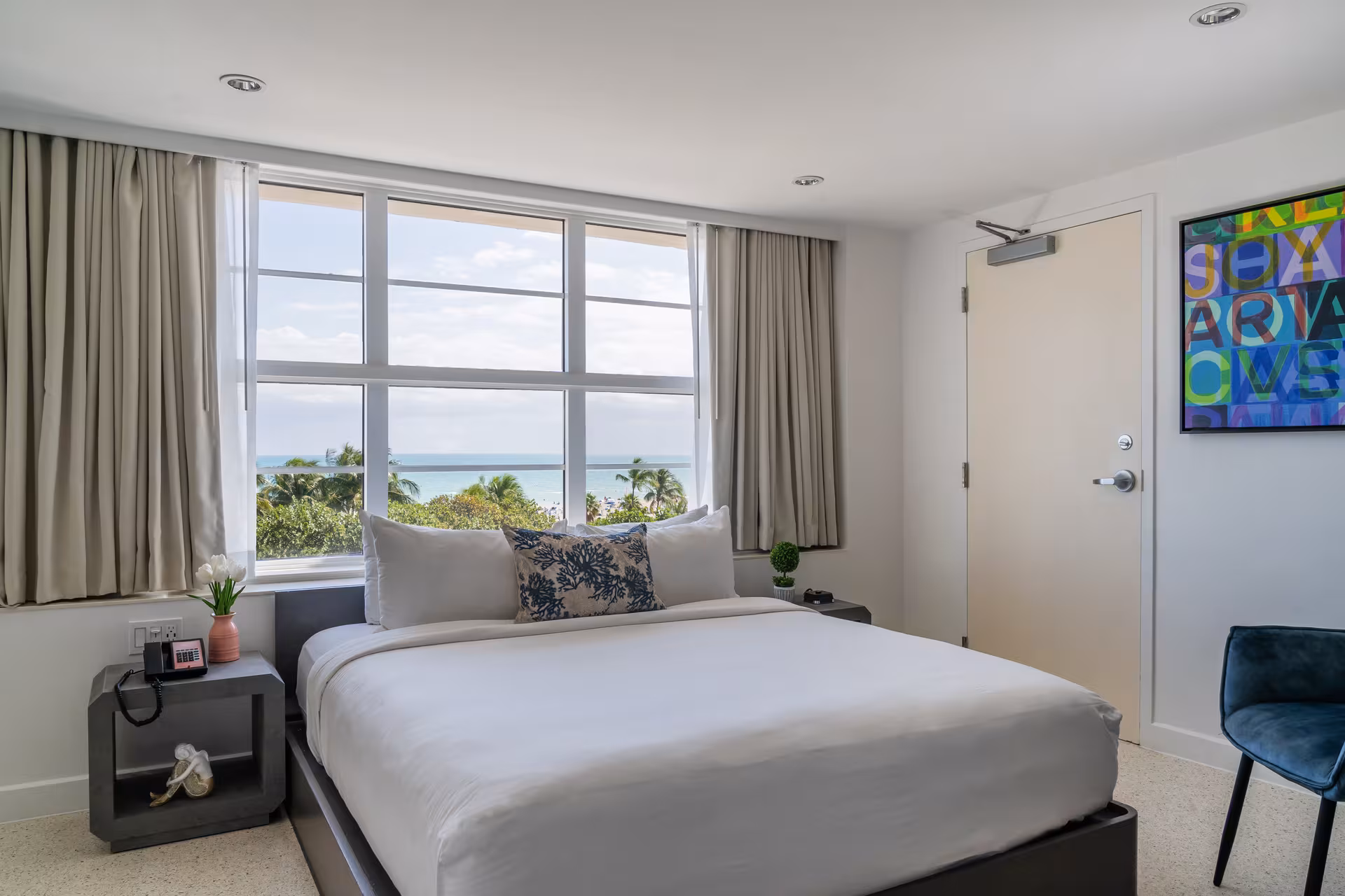 A wide-angle view of a hotel room with a large bed, nightstand, chair, and artwork on the wall.