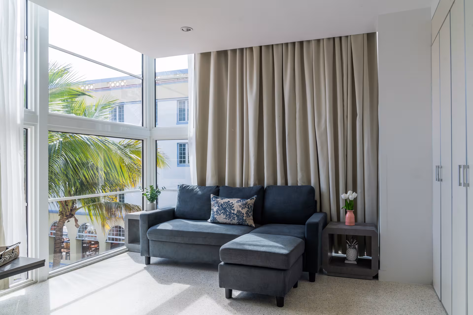 A living room with a gray sofa, ottoman, and a large window showing a palm tree outside.